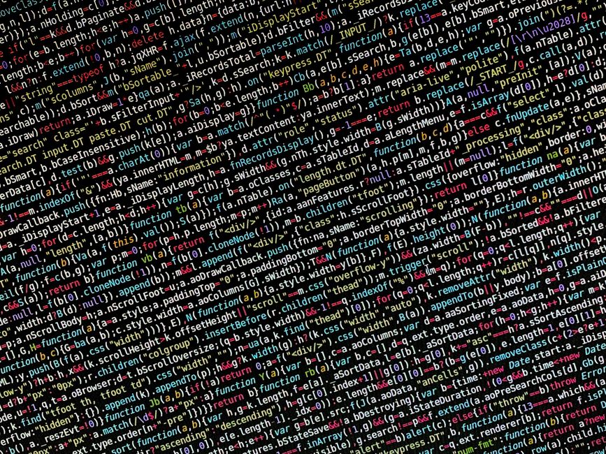 A close-up view of a silver laptop placed on a wooden table, displaying lines of code on its screen with a blurred background that appears to be an indoor workspace or home office. The laptop is open,